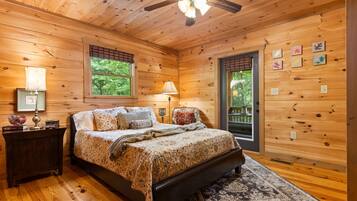 A wooden cabin bedroom with a bed, nightstands, a ceiling fan, and a view of the outdoors.