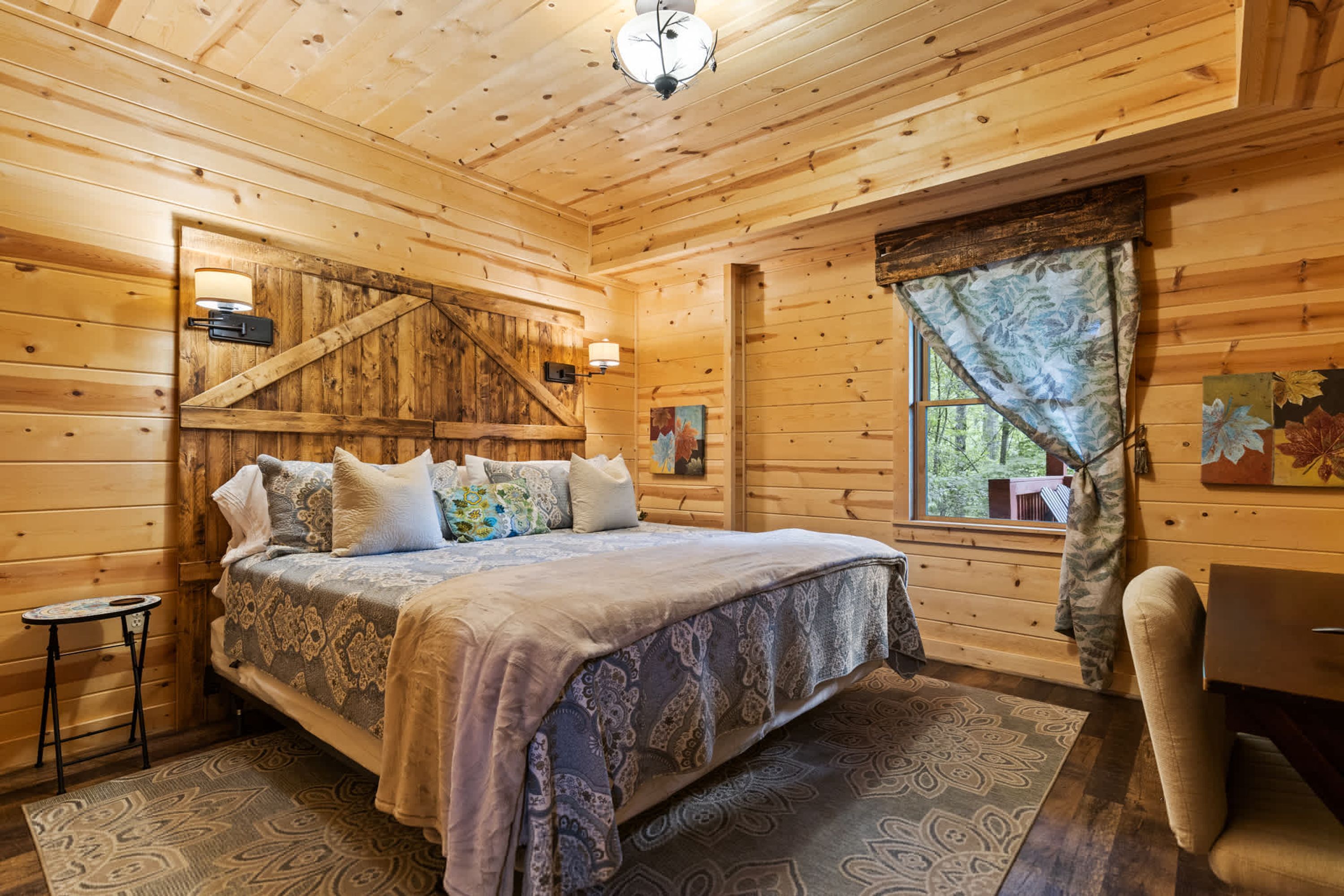 A wooden cabin bedroom with a bed, bedside tables, a window with curtains, and a wall-mounted lamp.