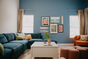 A living room with a sectional sofa, a white coffee table, and a small orange chair. There are two windows with curtains, and the walls are painted blue. - Sleeps 20 Games Basketball Pool Movie Room (Bradenton)