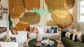 Ein Wohnzimmer mit moderner abstrakter Wandkunst, einem Neon-Schild mit der Aufschrift 'COCKTAILS & CACTI', einer Sofagarnitur, einem Couchtisch mit Snacks und einem Bücherregal mit Dekoration.