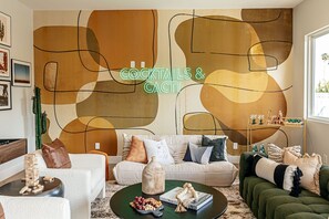A living room with a modern abstract wall art, a neon sign reading 'COCKTAILS & CACTI', a sofa set, a coffee table with snacks, and a bookshelf with decorative items. - A Desert Dream Perfect Getaway in Old Town (Scottsdale)