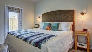 Ein Schlafzimmer mit Bett, Nachttisch, Wandlampe und einer Tür mit Blick ins Freie.