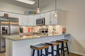 A kitchen with white cabinets, a stainless steel refrigerator, a microwave, a sink, and a granite countertop. There are three wooden bar stools and a hanging pendant light. - Borracho Beach- Waterslide Pet Friendly (Port Aransas)