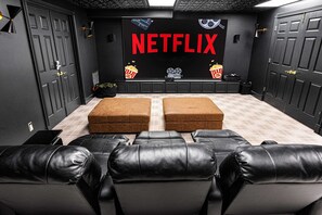 Sala de cinema com uma tela grande exibindo 'NETFLIX', dois pufes marrons e poltronas reclináveis de couro preto.