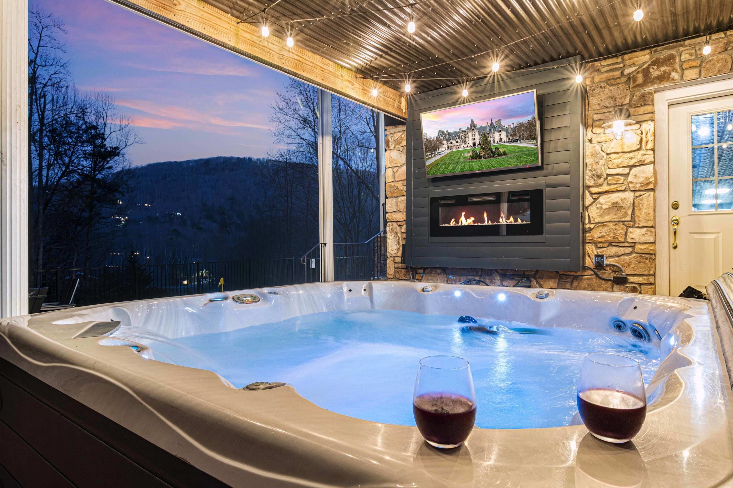 A hot tub with two glasses of wine, a fireplace, and a TV screen displaying a landscape.