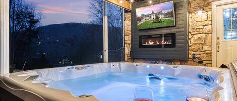 A hot tub with two glasses of wine, a fireplace, and a TV screen displaying a landscape.