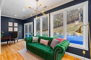 A room with a green sofa, a desk, framed pictures on the wall, and large windows with a view of a tennis court and trees. - 7k Sqft Estate - Epic Amenities Fun - Sleeps 20 (Asheville)