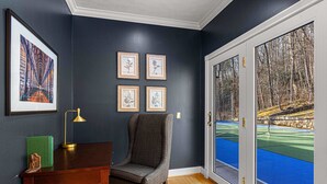 Una stanza con una parete blu scuro, una scrivania in legno, una sedia, un'illustrazione botanica incorniciata e un'ampia finestra che si affaccia su un campo da tennis blu e alberi.