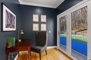 A room with a dark blue wall, a wooden desk, a chair, a framed botanical illustration, and a large window with a view of a blue tennis court and trees. - 7k Sqft Estate - Epic Amenities Fun - Sleeps 20 (Asheville)