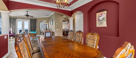 A dining room with a large wooden table and ornate chairs, a chandelier, and a view of the living area.