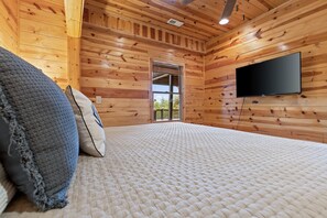 Une pièce aux murs en bois, un lit recouvert d’une courtepointe blanche à motifs, un téléviseur à écran plat fixé au mur et une porte donnant sur l’extérieur.