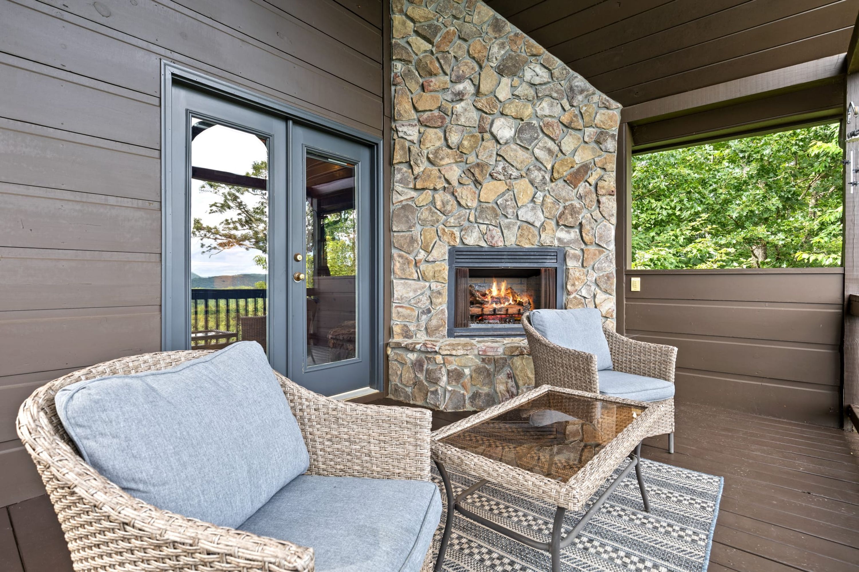 A covered outdoor seating area with wicker furniture, a stone fireplace, and a view of greenery.