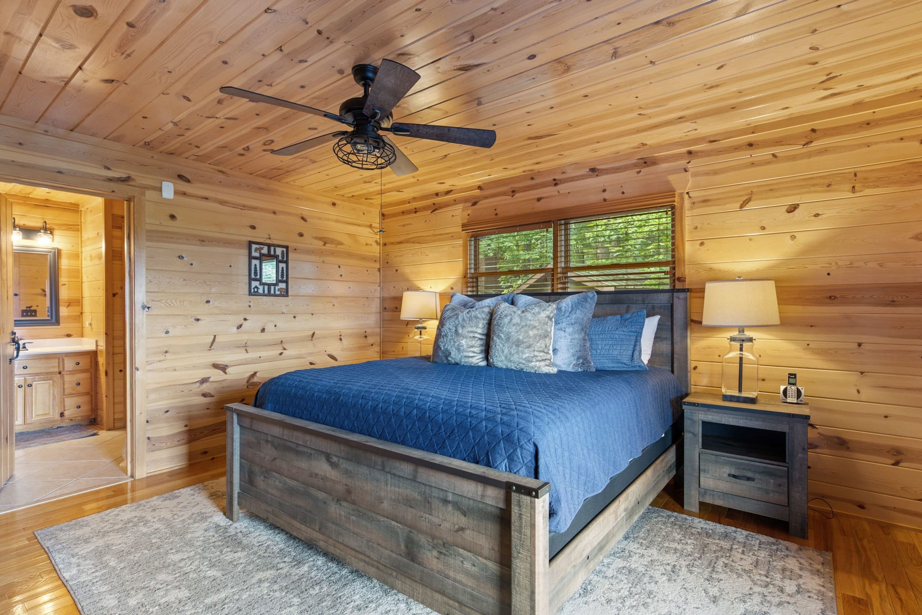 Une chambre dans une cabane en bois, avec un lit, deux lampes de chevet, un ventilateur de plafond et une fenêtre donnant sur les arbres.