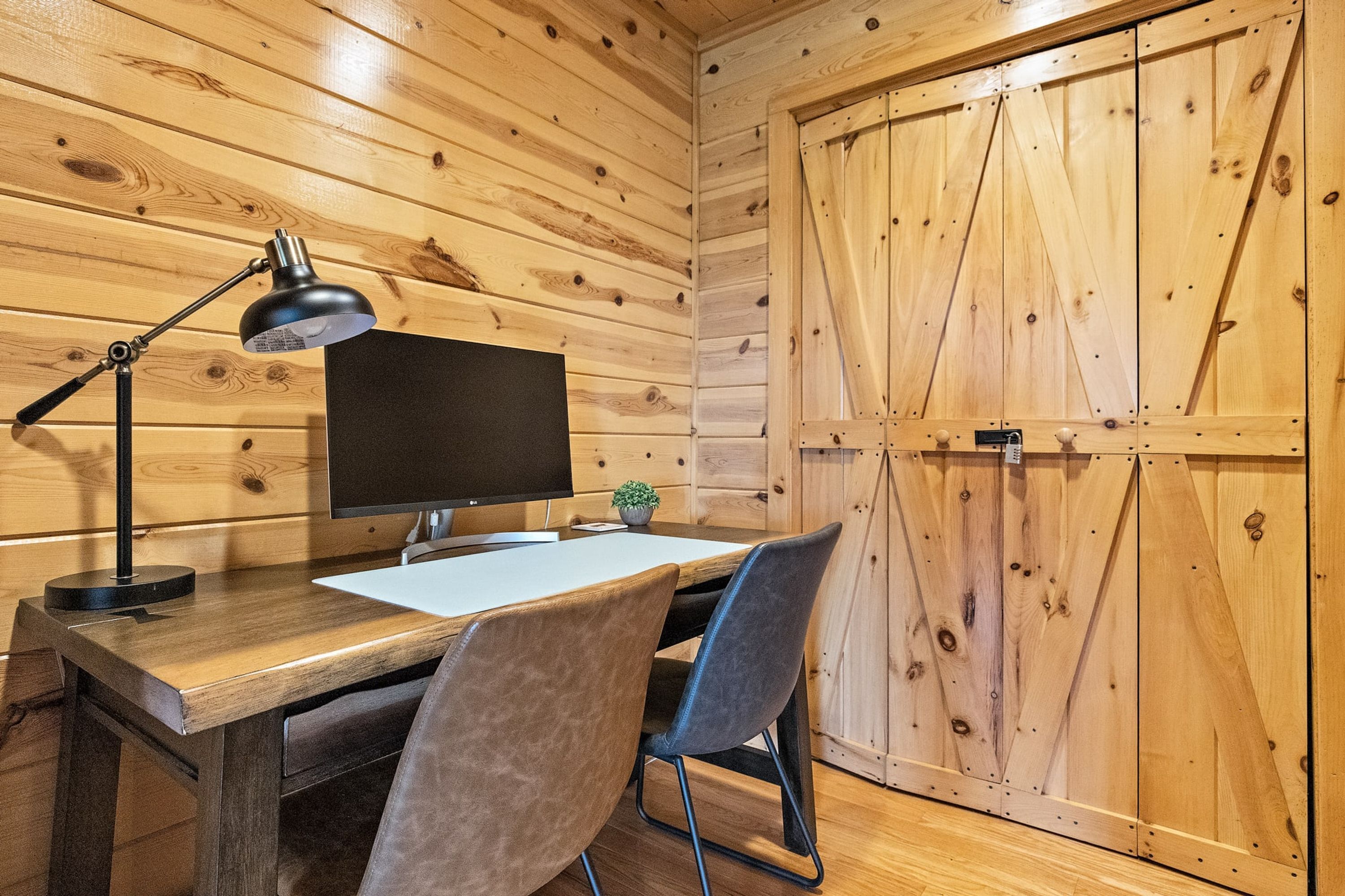 Interior de madera con un escritorio, silla, monitor de computadora, lámpara y una puerta de madera cerrada.