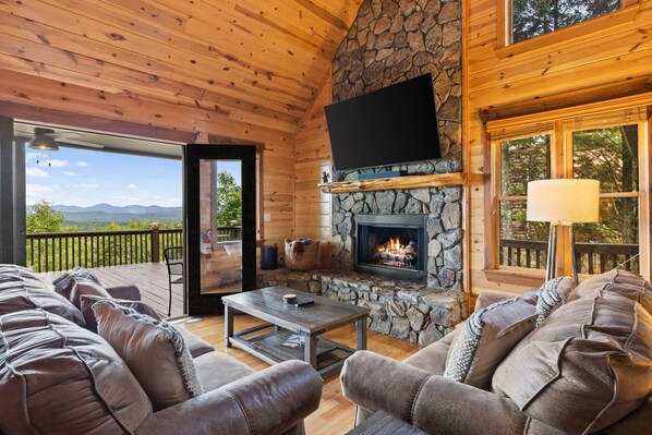 L’intérieur chaleureux d’un chalet avec une cheminée en pierre, un plafond en bois et une grande fenêtre donnant sur un paysage pittoresque.