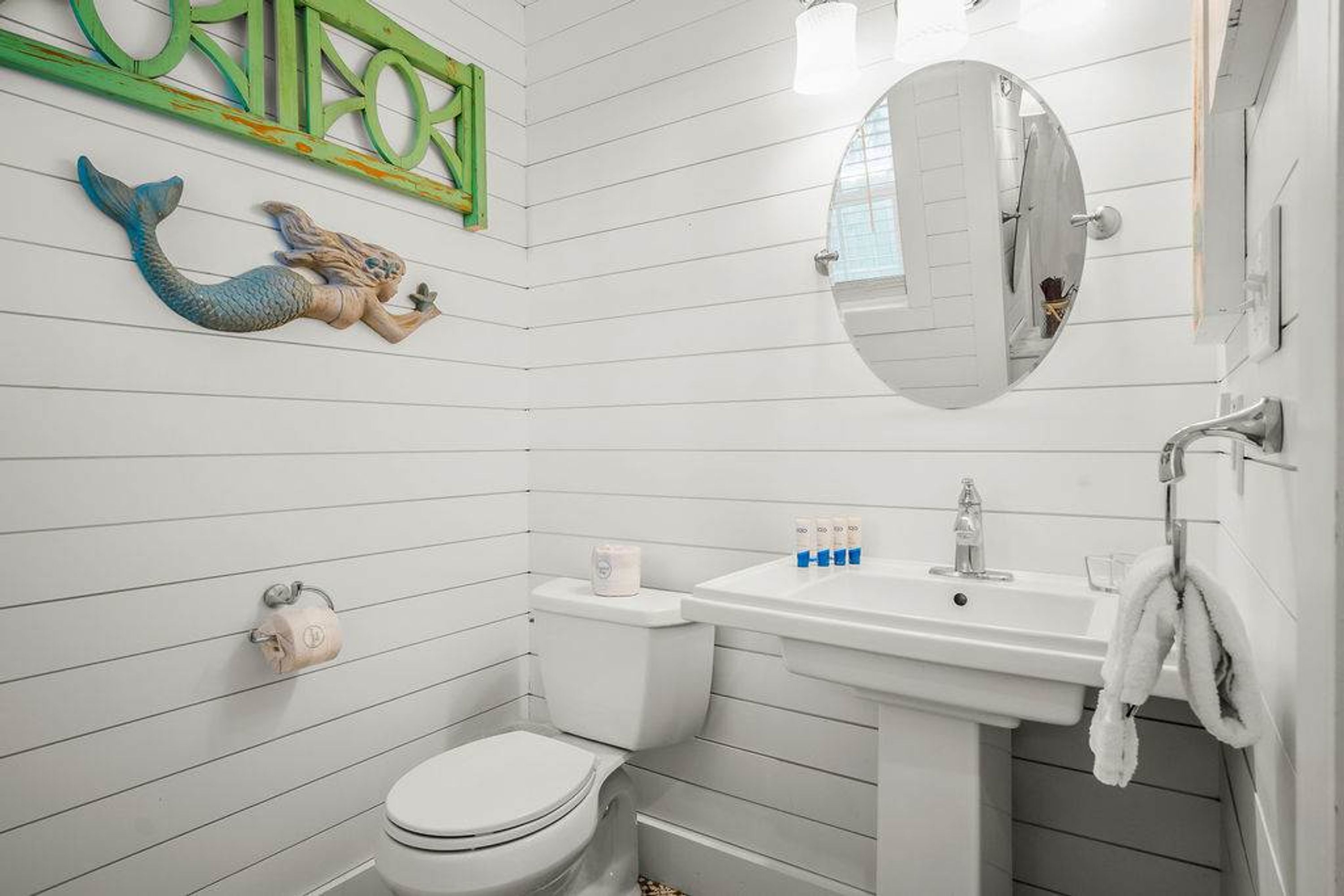 Ein Badezimmer mit weiß gefliesten Wänden, einer Toilette, einem Waschbecken mit Pflegeprodukten und einem Spiegel.