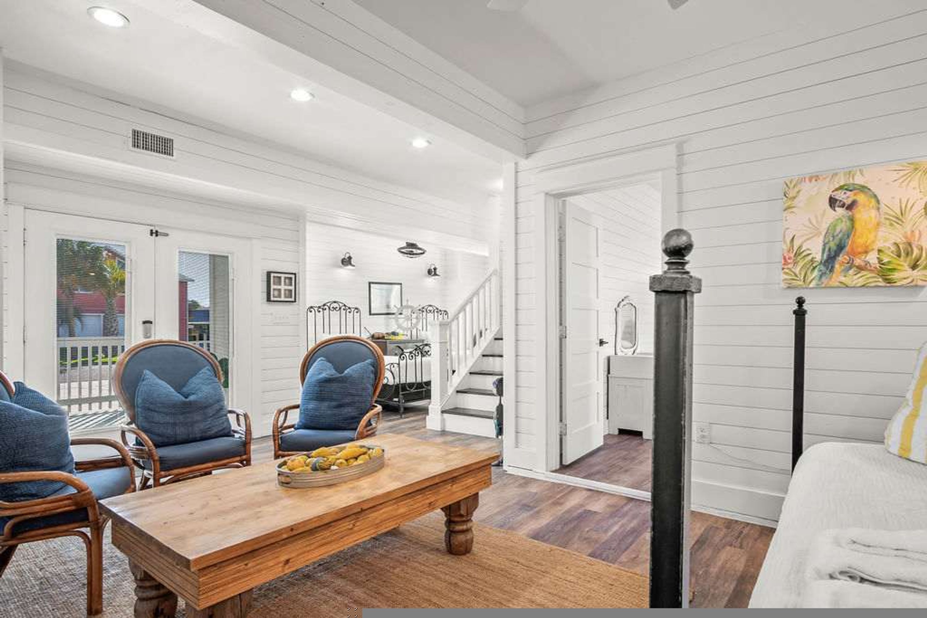 A spacious living room with a wooden coffee table, two blue upholstered chairs, and a white sofa. A staircase leads to an upper level, and a painting of a parrot is visible on the wall.