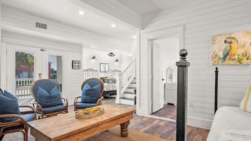 A spacious living room with a wooden coffee table, two blue upholstered chairs, and a white sofa. A staircase leads to an upper level, and a painting of a parrot is visible on the wall.