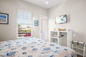 Um quarto com cama, janela com persianas, porta, prateleira com toalhas e um quadro na parede.