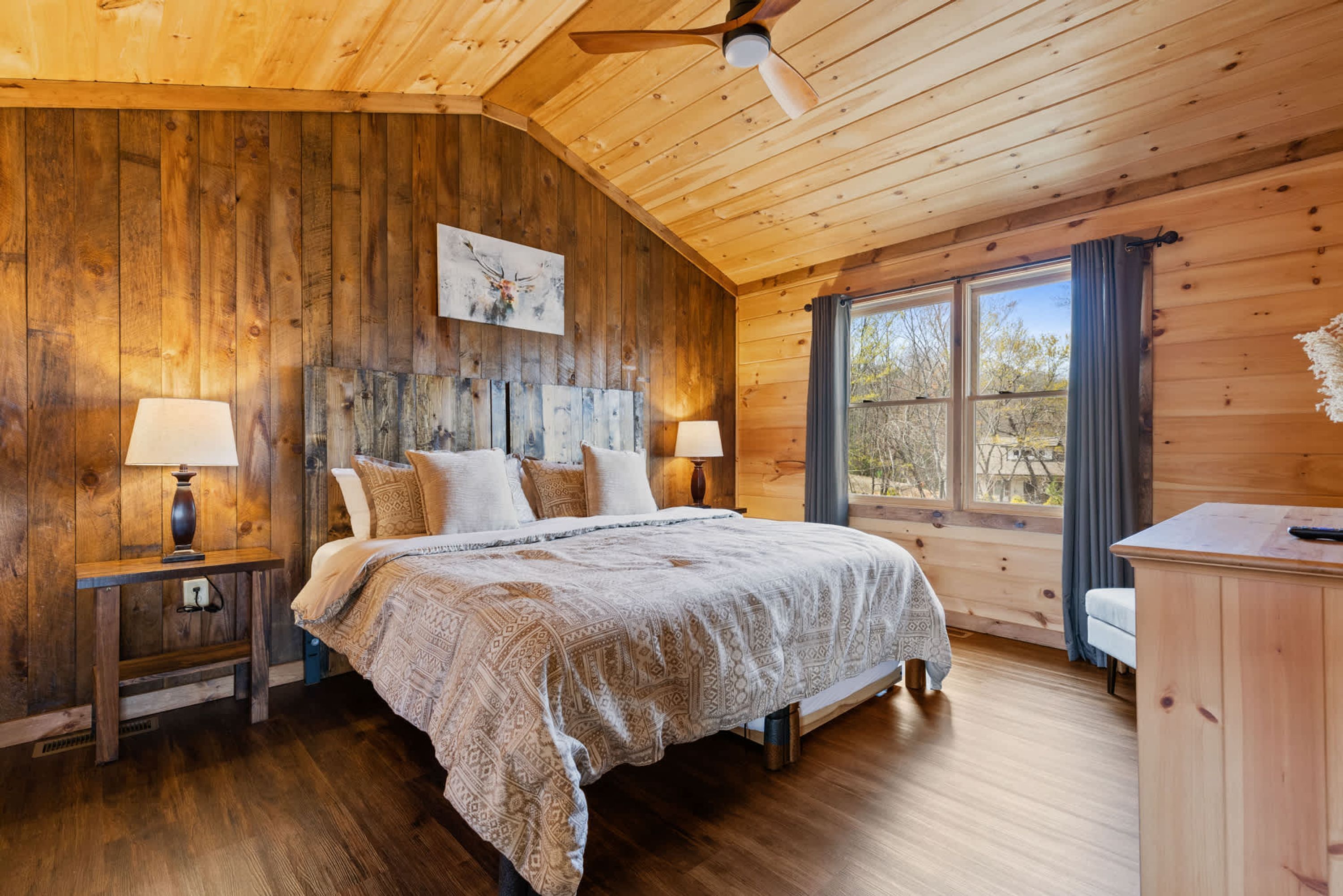 A wooden cabin bedroom with a bed, two nightstands, a ceiling fan, and a large window with curtains.