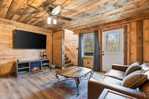 Interior de una cabaña de madera con chimenea, televisor montado en la pared, sofá y una escalera que conduce a un nivel superior.