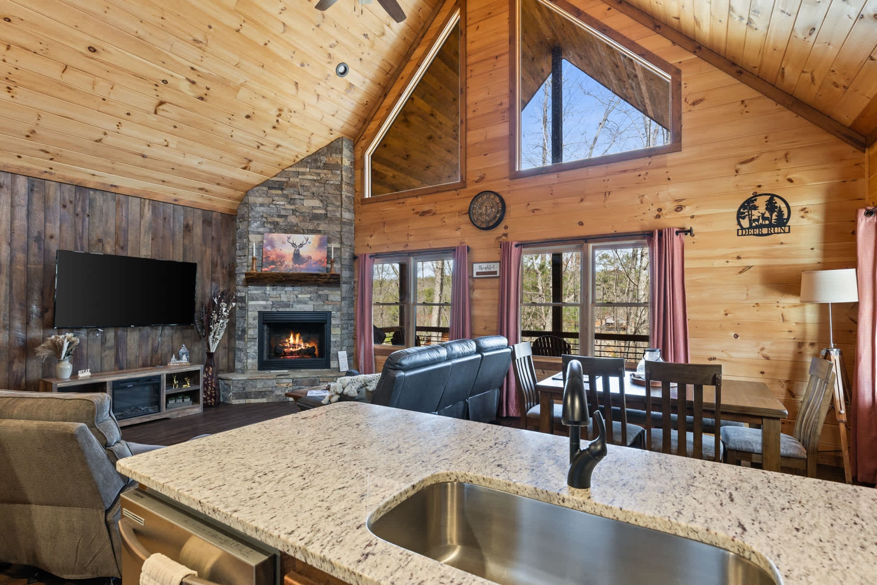A spacious living area with a stone fireplace, a flat-screen TV mounted on the wall, and a large window offering a view of the outdoors.