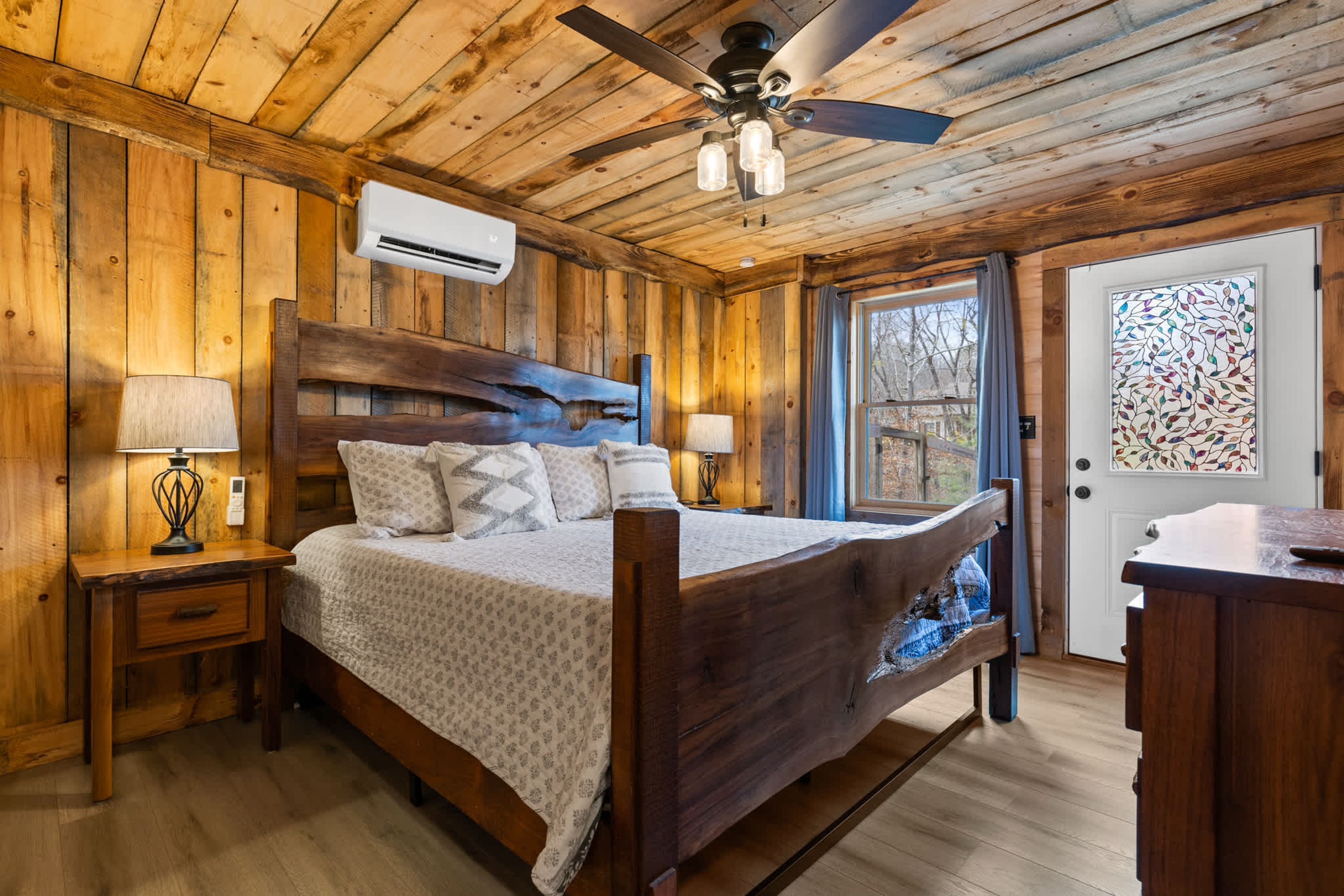 A wooden cabin bedroom with a bed, bedside lamps, a ceiling fan, and a door with a stained glass window.