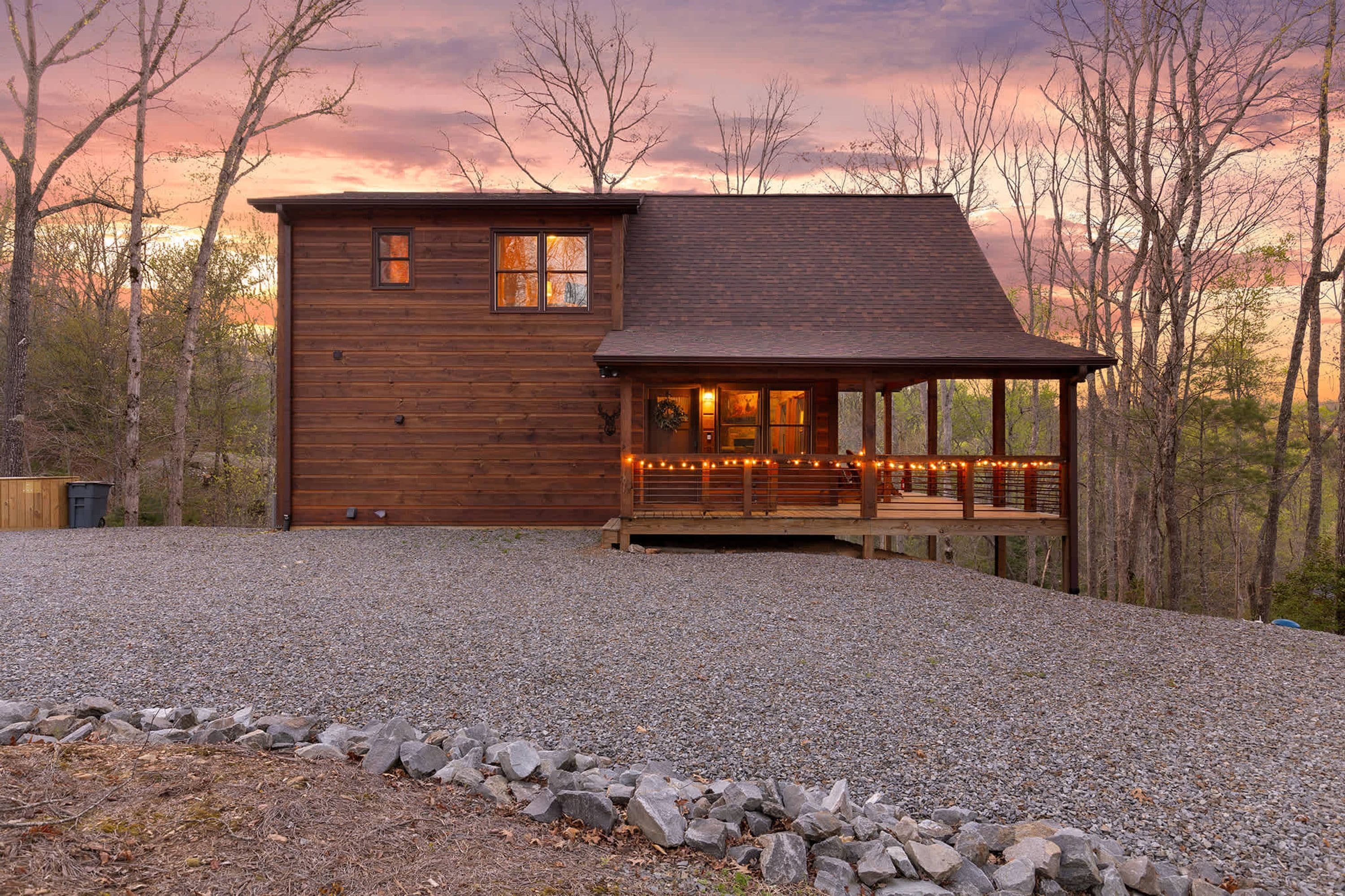 A wooden cabin with a porch, surrounded by trees and a gravel driveway.
