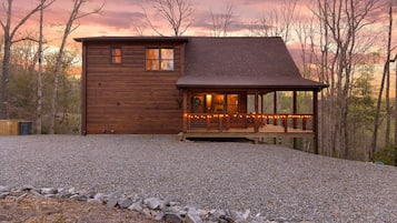 A wooden cabin with a porch, surrounded by trees and a gravel driveway.