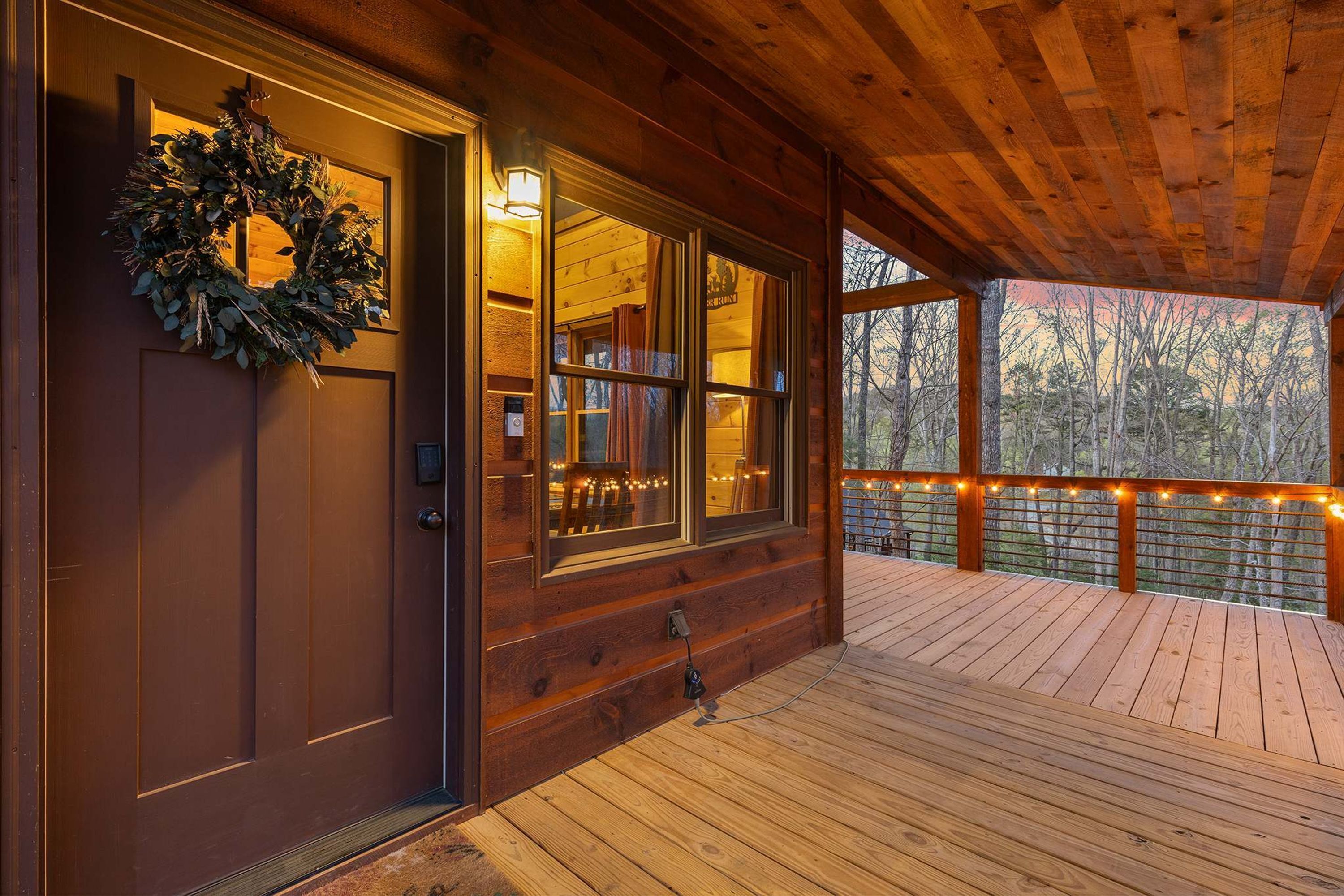 A wooden cabin with a porch, a wreath on the door, and string lights on the railing.
