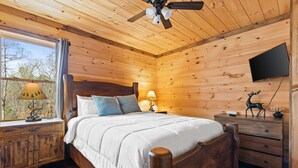 Een slaapkamer in een houten chalet, met een bed, nachtlampen, een televisie en een raam met uitzicht op bomen.
