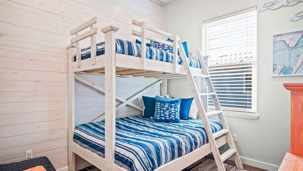 Een stapelbed met blauw beddengoed, een ladder en een raam met zonwering.