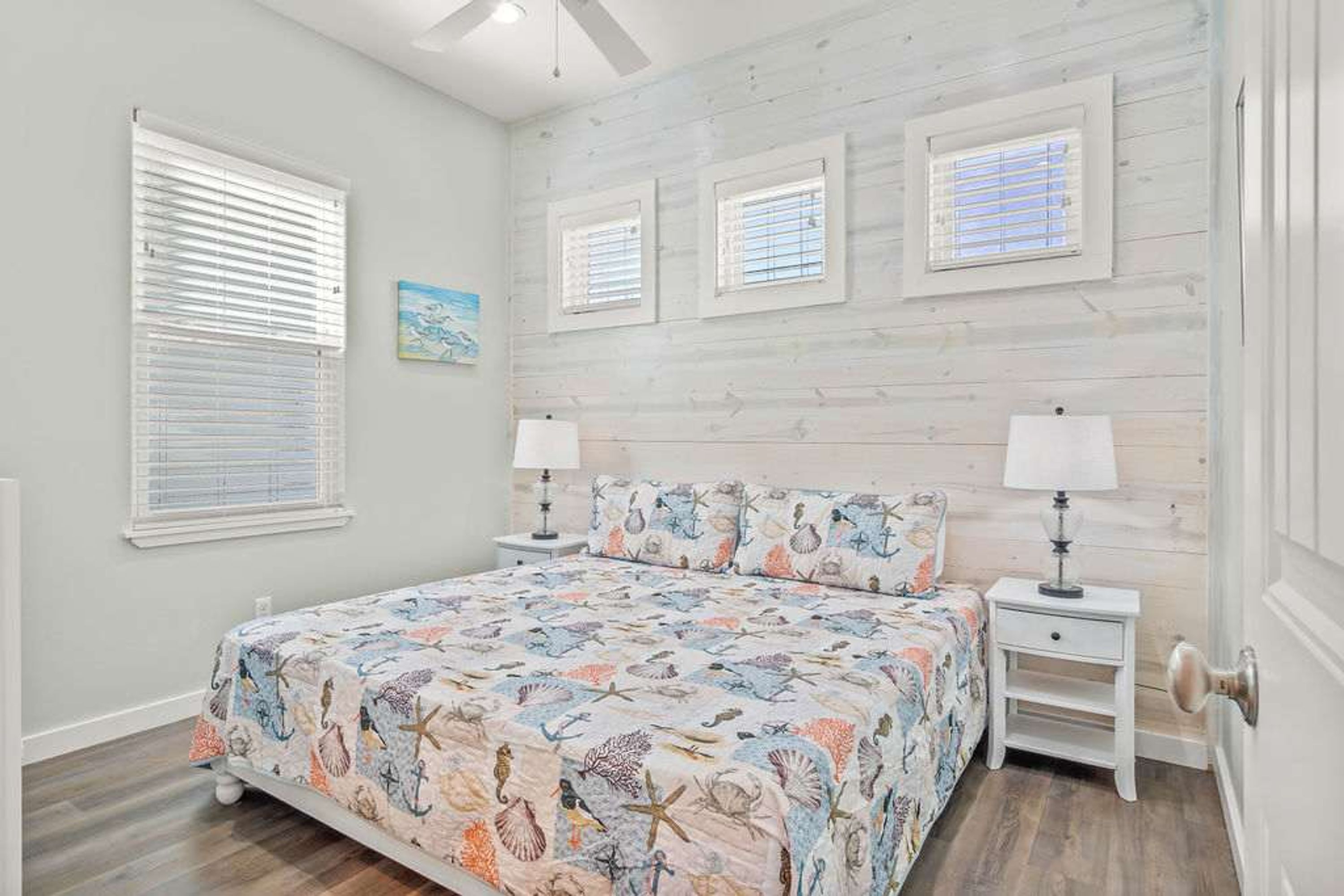 A bedroom with a bed, two bedside tables, a ceiling fan, and three windows with blinds.