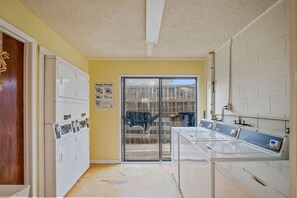 A laundry facility with multiple washing machines and dryers, a glass door leading outside, and a poster on the wall. - The Codfather- Family Friendly 2 Minute to Beach (Port Aransas)