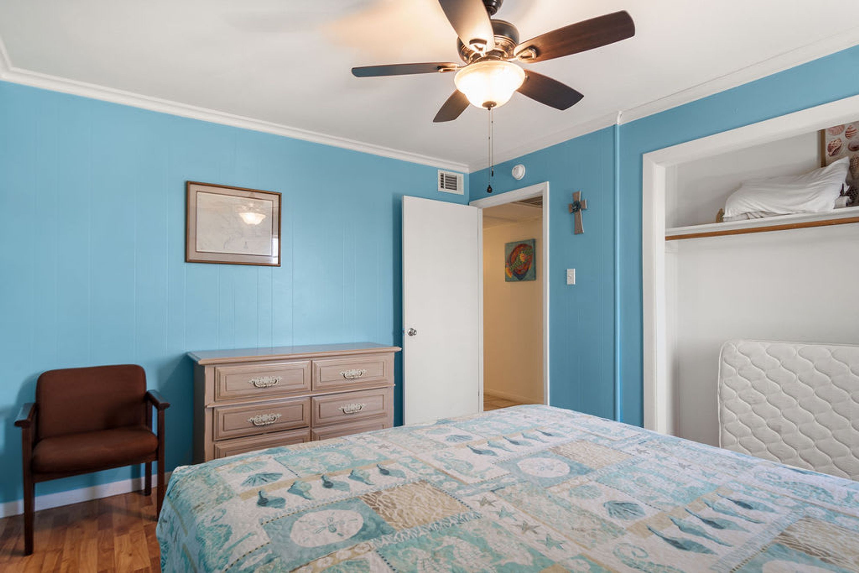 Un dormitorio con cama, cómoda, silla, ventilador de techo y una puerta que da a otra habitación.