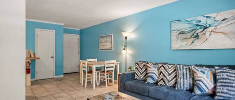 A living room with a blue wall, a sofa with patterned cushions, a dining table with chairs, and a floor lamp.