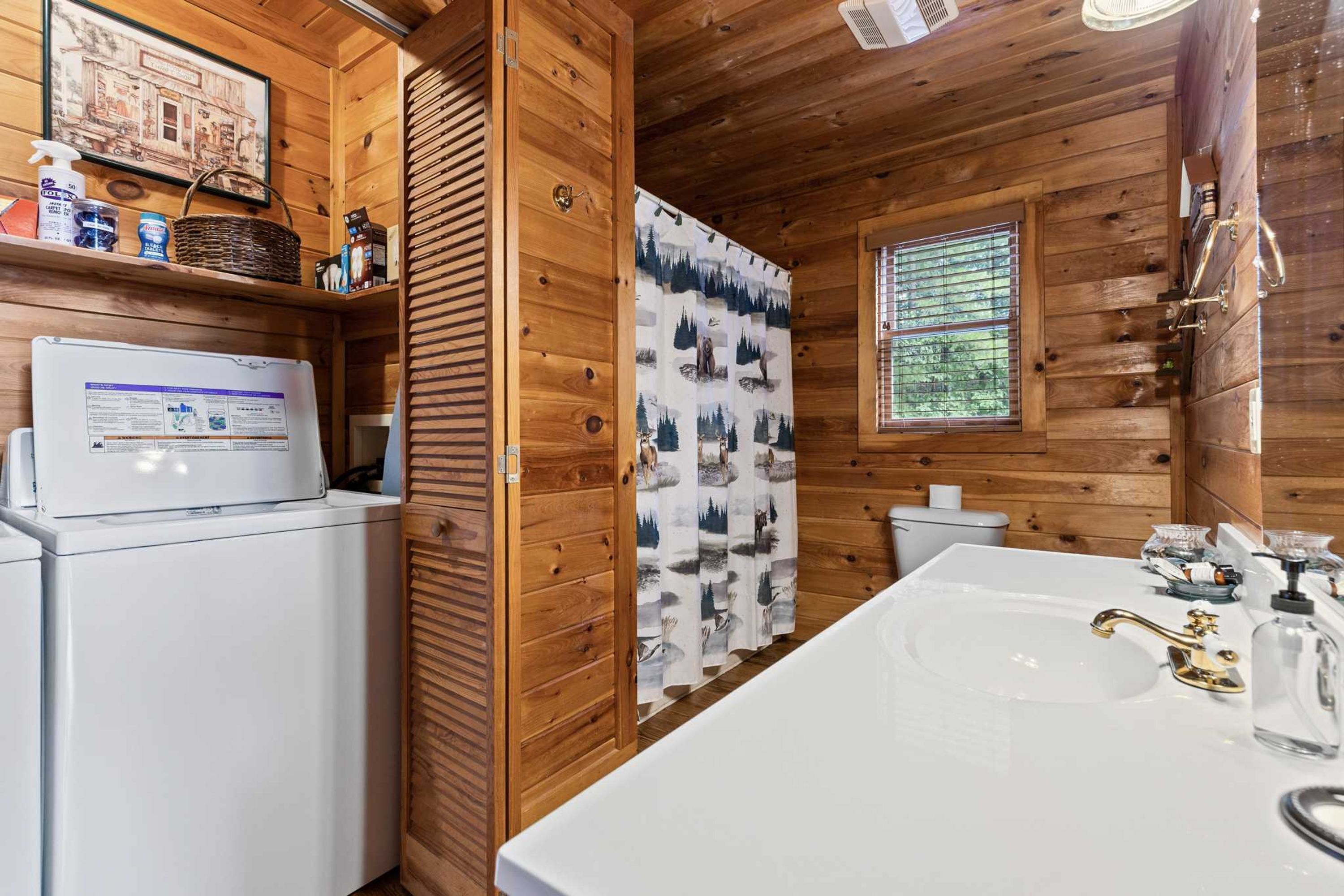Interieur van een houten hutje met een wasmachine, een spoelbak met kraan, een raam met zonwering en een wand met een decoratief gordijn.