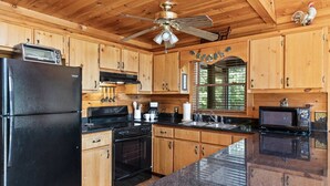 A kitchen with wooden cabinets, a black refrigerator, a stove, a microwave, and a ceiling fan. - Mountain Views-hot Tub-games-pool Foosball (Morganton)