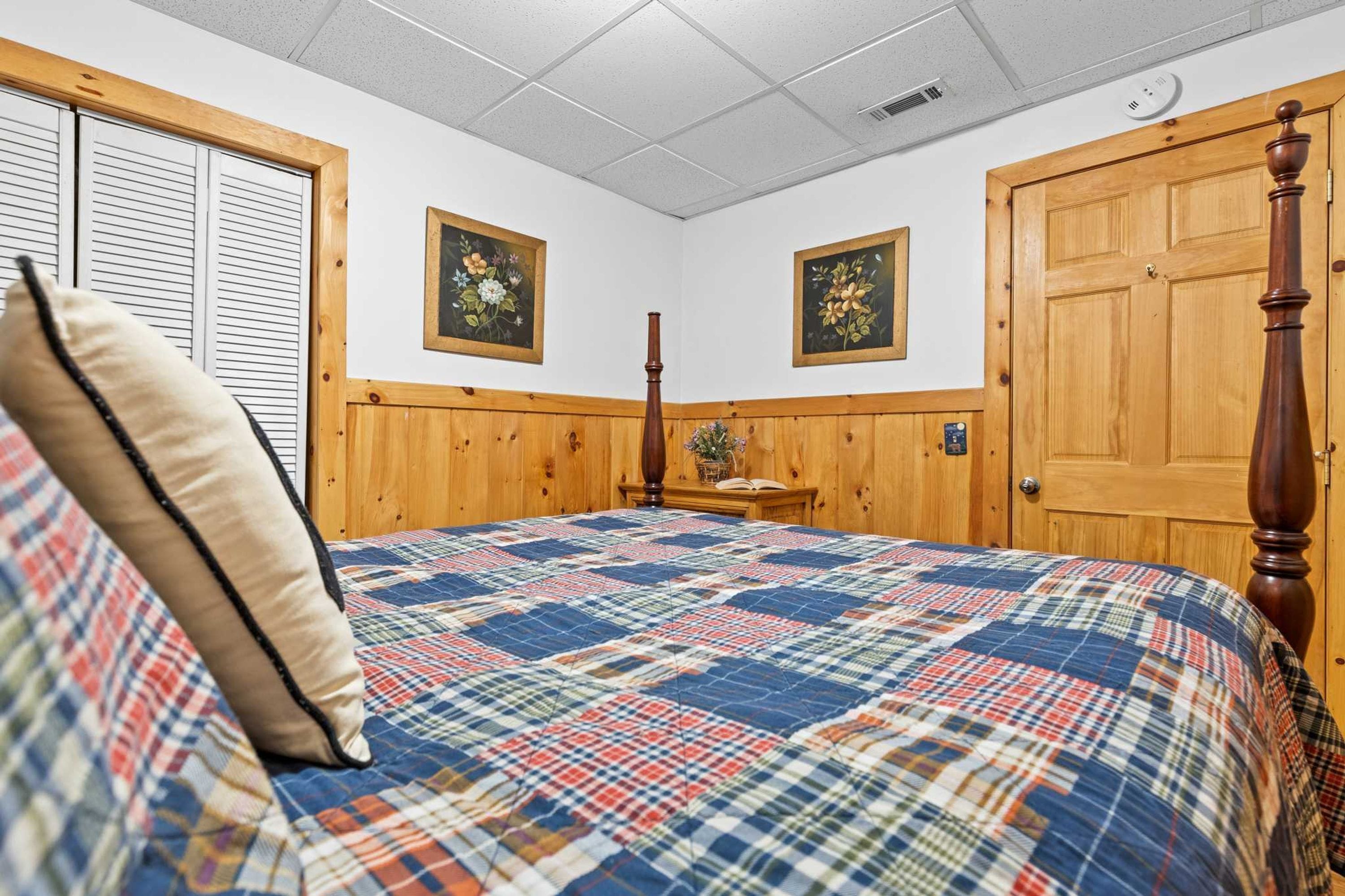 Un dormitorio con cama, paneles de madera, dos cuadros en la pared, una puerta y una ventana con persianas.