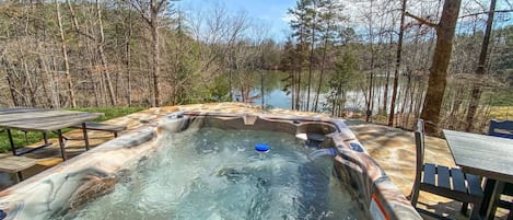 A hot tub surrounded by a wooden deck with benches, set against a backdrop of trees and a body of water.
