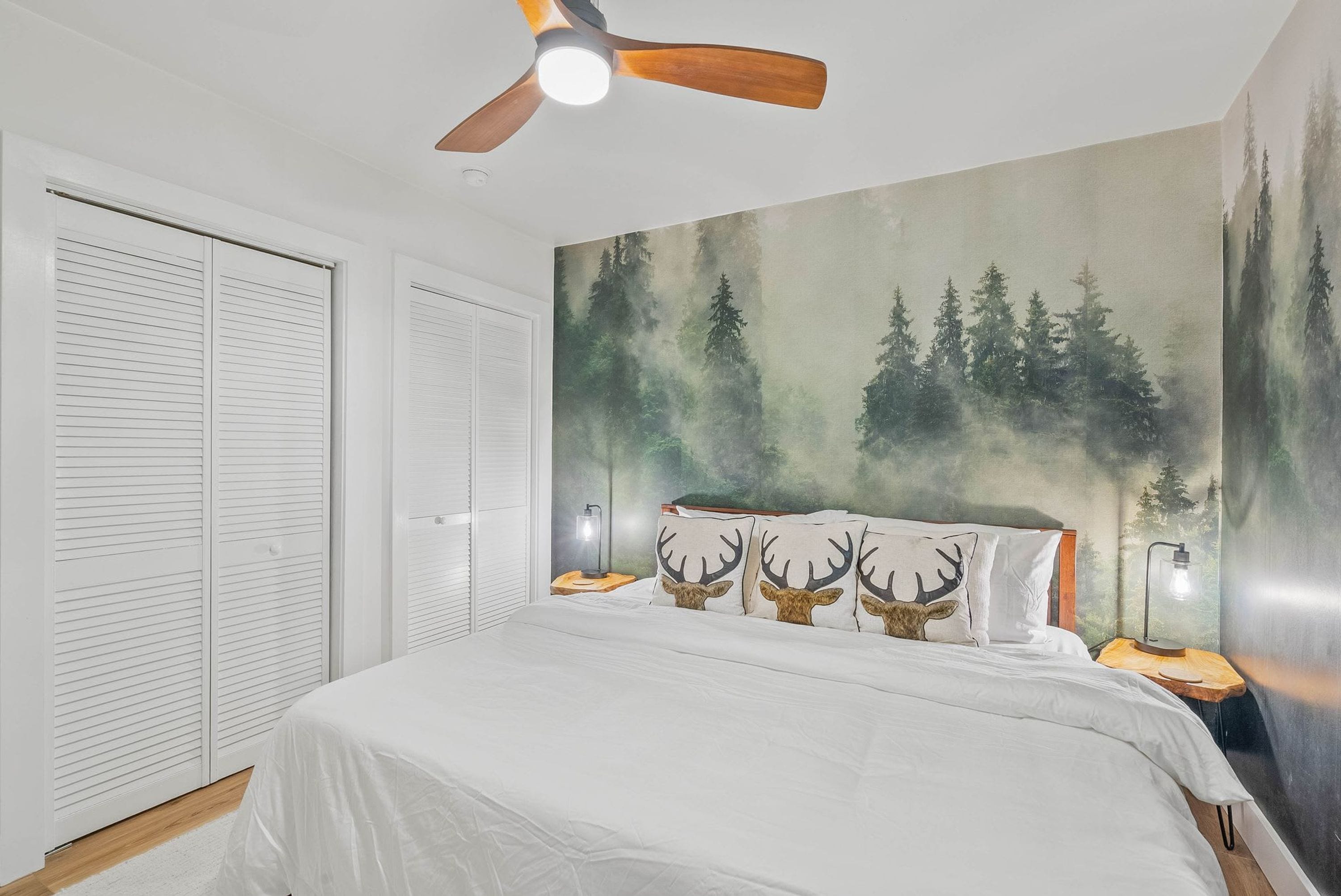 A bedroom with a bed, white curtains, a ceiling fan, a forest mural, and two bedside lamps.
