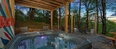 Un espace extérieur aménagé avec un jacuzzi et une pergola en bois, entouré d’arbres et doté de sièges extérieurs.