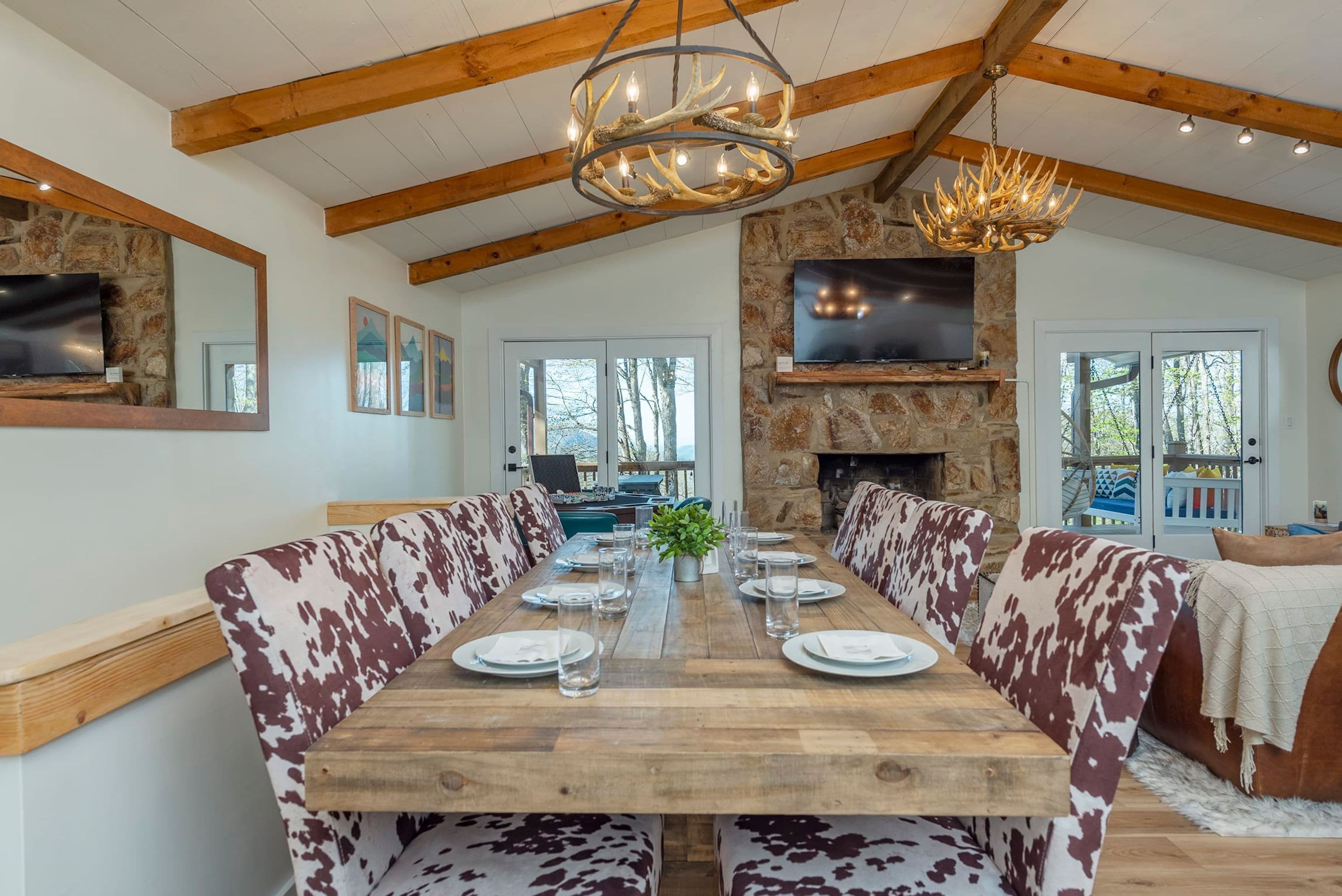 A dining area with a long wooden table set for four, flanked by patterned sofas. A stone fireplace and a mounted television are visible in the background.