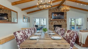 A dining area with a long wooden table set for four, flanked by patterned sofas. A stone fireplace and a mounted television are visible in the background.