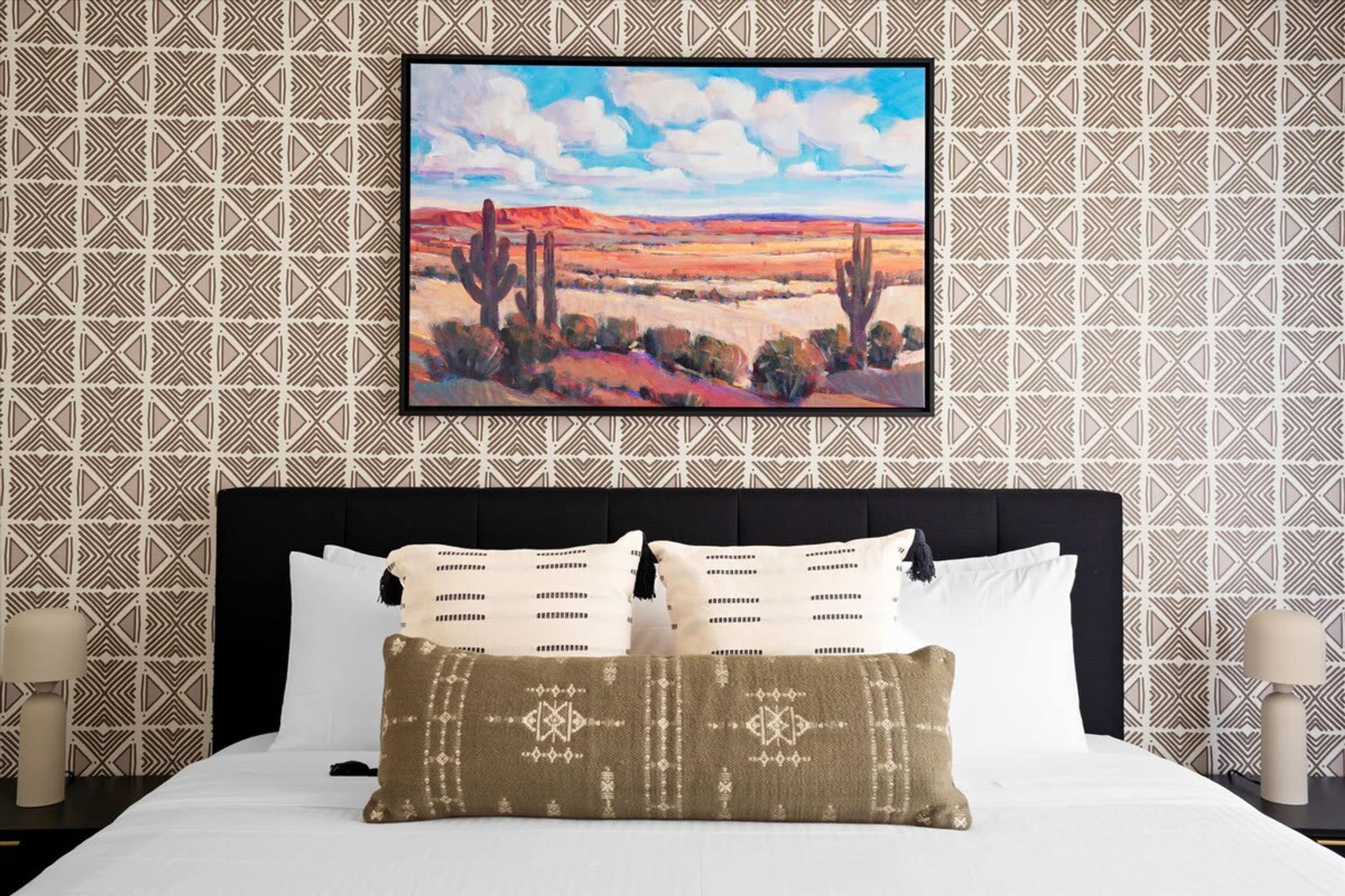 Ein Hotelzimmer mit einem großen Bett, einem gerahmten Gemälde einer Wüstenlandschaft und Wandtapete mit Muster.