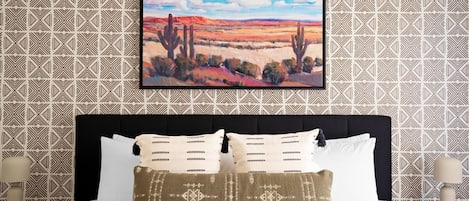 A hotel room with a large bed, a framed painting of a desert landscape, and patterned wallpaper.