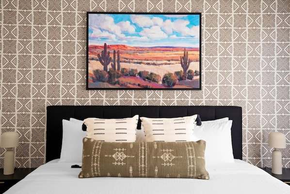 A hotel room with a large bed, a framed painting of a desert landscape, and patterned wallpaper. - Family Fun Sleeps 12 Pool Fire Pit Puttputt (Oro Valley)
