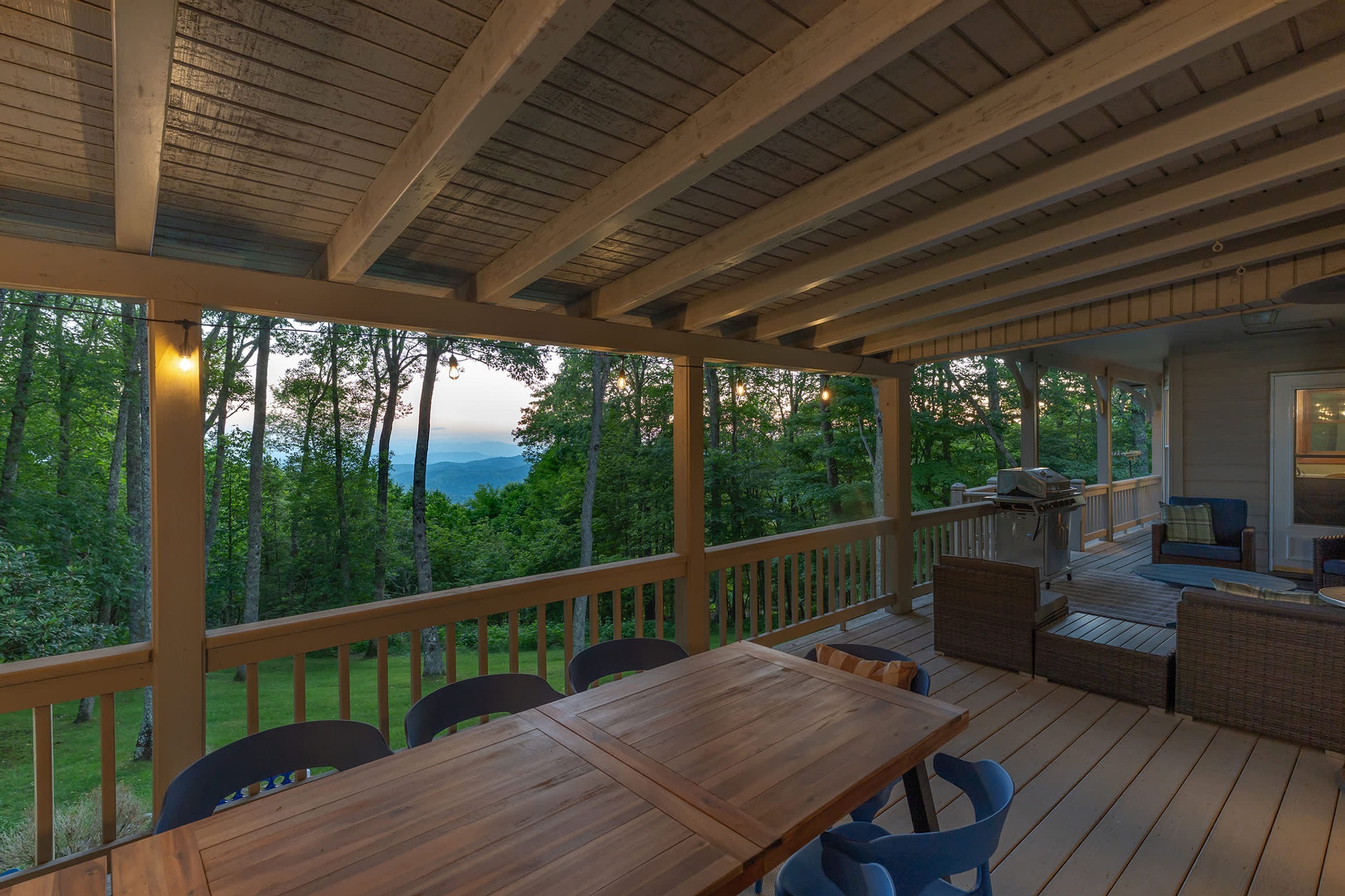 A covered outdoor deck with a wooden table and chairs, overlooking a forested landscape.