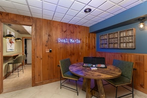 Um quarto com paredes de painel de madeira, um letreiro de neon com a inscrição 'Deal Me In', uma mesa de madeira redonda com cadeiras e um aparelho de iluminação fixado na parede.