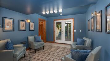 A room with blue walls, a tiled floor, and framed botanical prints. There are two leather sofas with blue pillows, a wooden door, and a glass door leading outside.